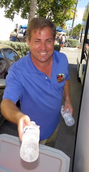 Man in a blue polo shirt smiling and handing out bottled water outdoors near parked vehicles and trees.