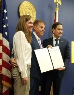 Three people standing in front of a U.S. flag and a seal, holding an award certificate.