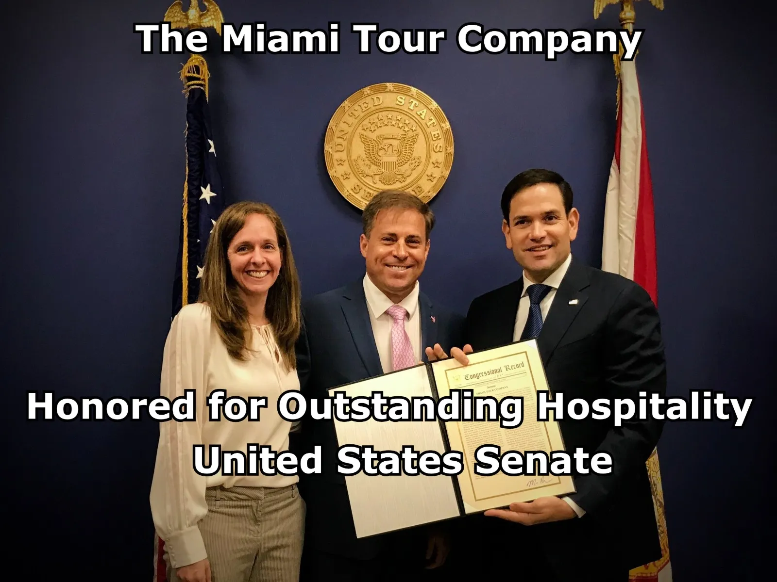 Three people standing and smiling in front of United States Senate seal and flags, holding a certificate of recognition for The Miami Tour Company.