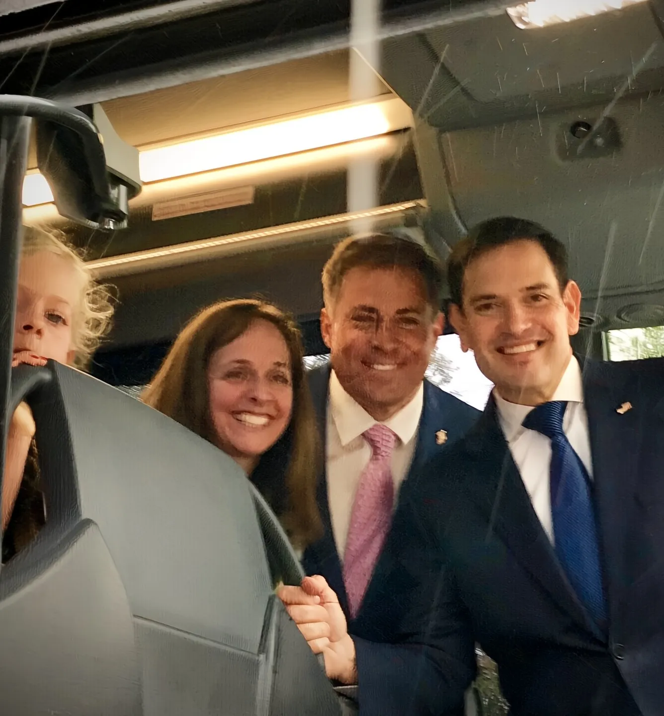 Four people smiling inside a vehicle, including a child peeking over a car seat and three adults dressed formally.