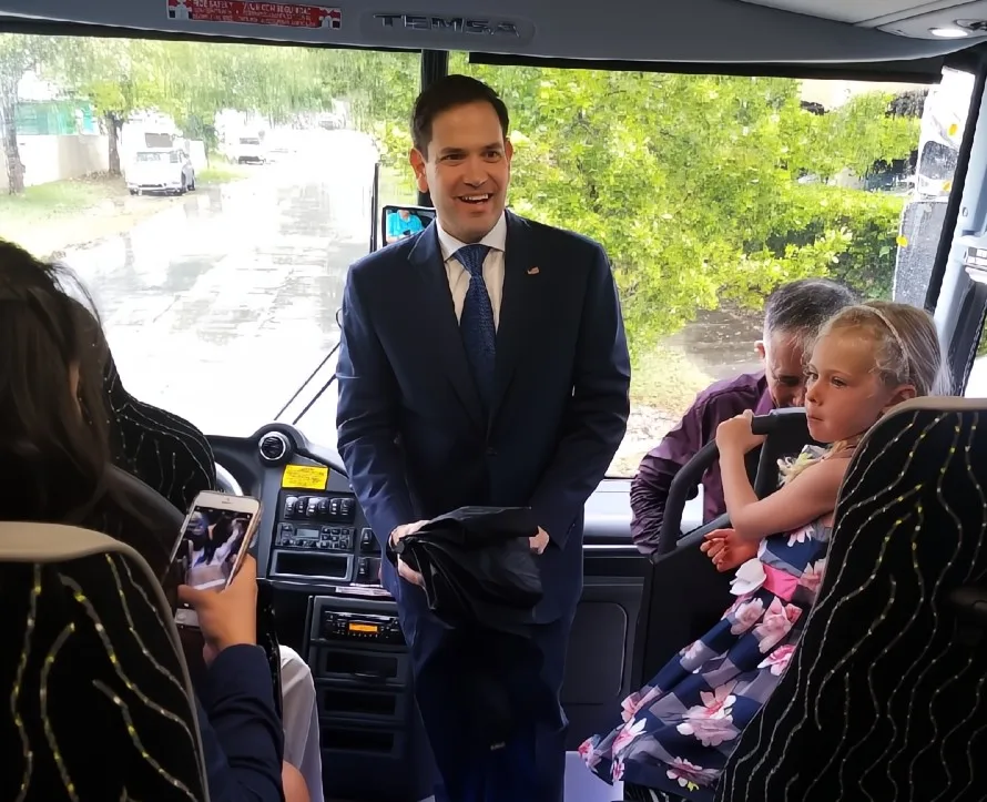 Man in a navy suit smiling inside a bus with a young girl in a floral dress sitting and a person taking a photo with a smartphone.