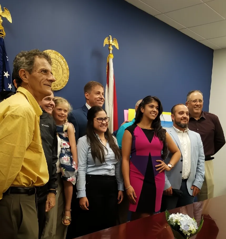 A diverse group of people smiling and posing together in a room with blue walls, U.S. and state flags, and an official seal.