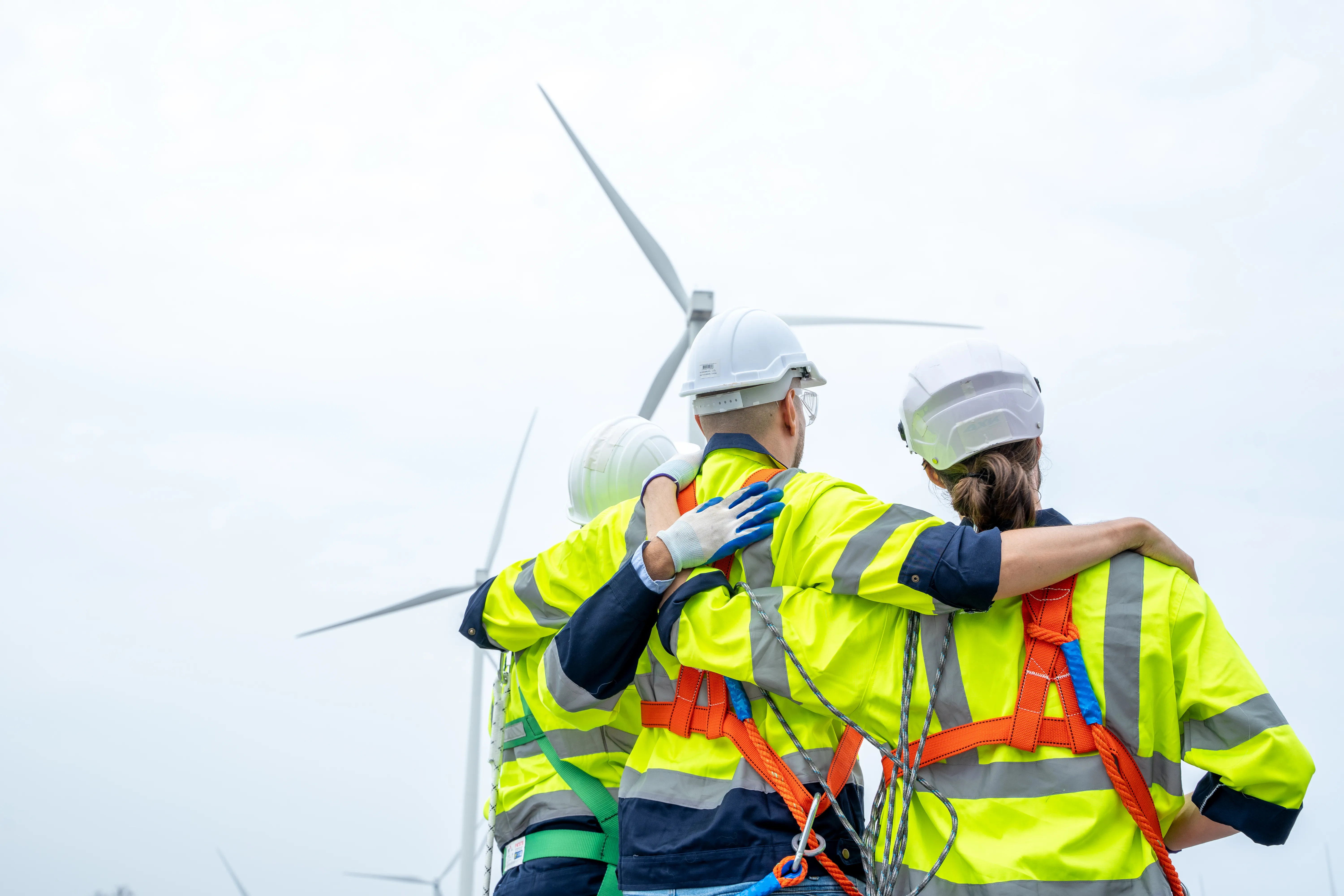 Three engineers in high-visibility jackets and safety helmets with arms around each other, standing in front of wind turbines.