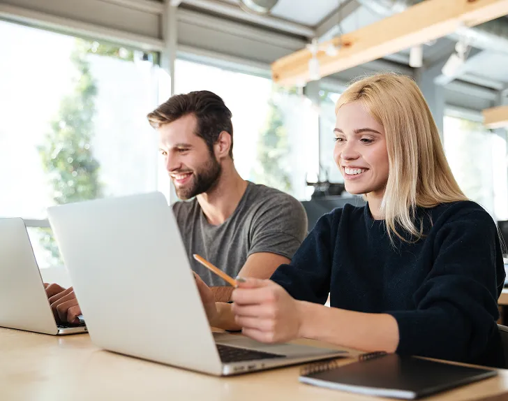 Two colleagues, a man and a woman, smiling as they work together on laptops in a bright office.