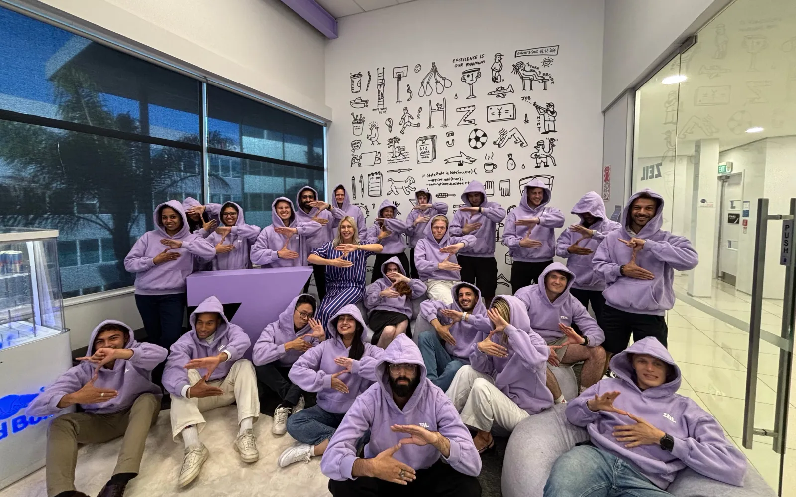 Group of people wearing matching lavender hoodies posing indoors with hands forming a 'Z' shape, in front of a white wall with black doodles.