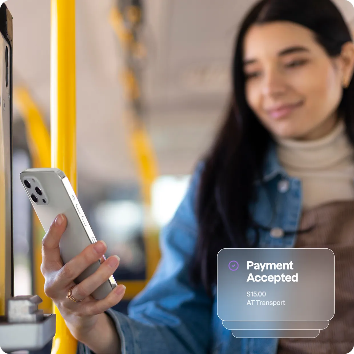 Woman in denim jacket holding phone near a yellow pole on public transport with a payment accepted notification for $15.