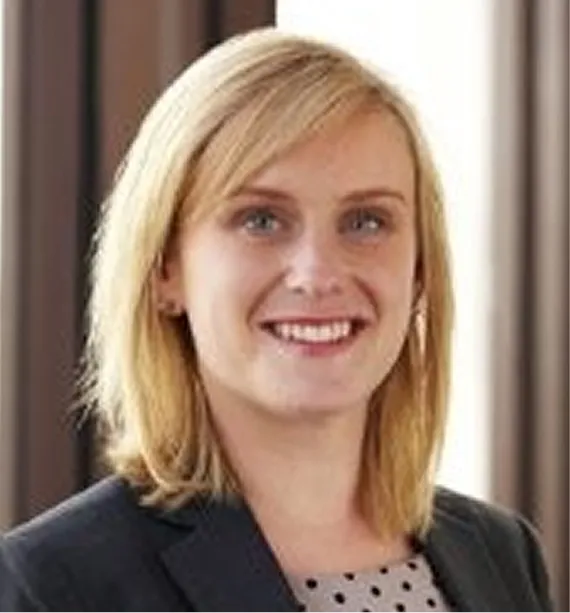 Smiling woman with blonde hair wearing a dark blazer and a polka dot top in an indoor setting.