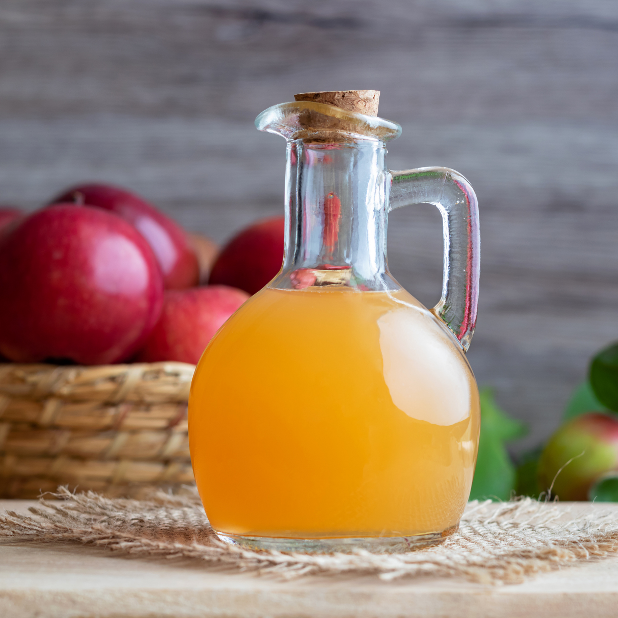 A glass jug filled with amber colored apple cider vinegar, with a basket of red apples in the background.