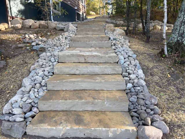 Outdoor stone steps bordered by gray and brown rocks leading uphill beside trees and a wooden building.