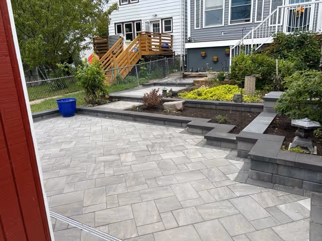 Backyard with newly installed gray stone paver patio, tiered garden beds with plants, and wooden stairs leading to a house.