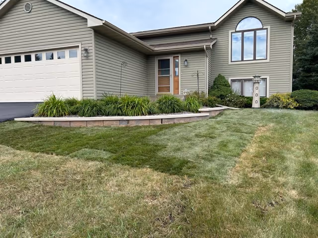 Front view of a beige suburban house with a two-car garage, a small raised garden bed with green plants, and a lawn with patches of uneven grass.
