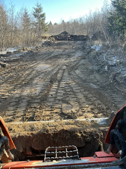 View from a bulldozer showing freshly leveled dirt road with tire tracks and trees on both sides.