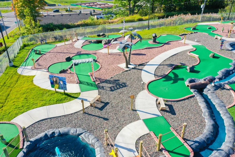 Aerial view of a colorful mini golf course with multiple putting greens, pathways, and decorative obstacles including palm trees and a water fountain.