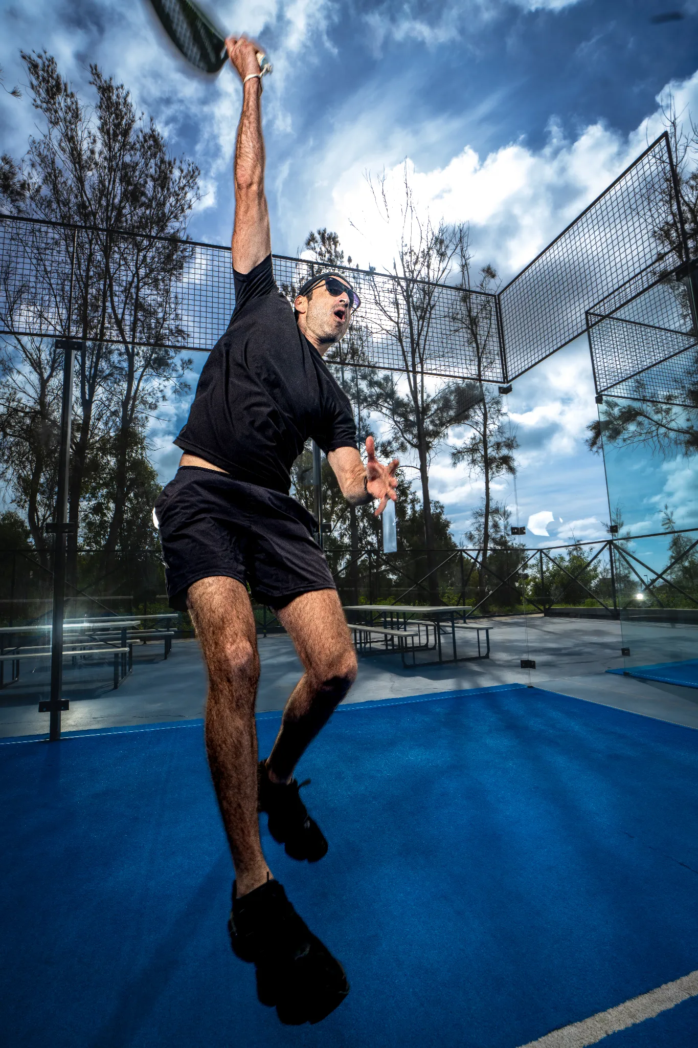 Hombre golpeando una pelota en una cancha de pádel con ropa deportiva negra y gafas de sol.