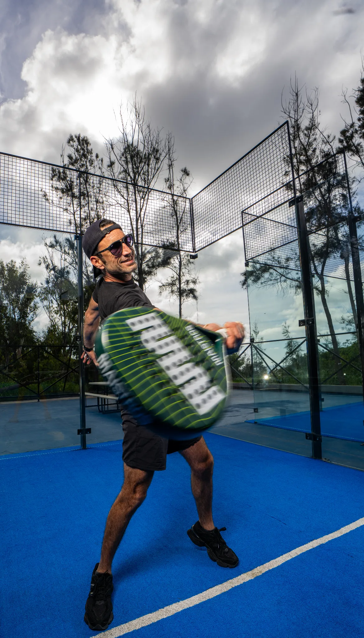 Hombre jugando pádel en cancha azul con raqueta verde y negra, lleva gorra y gafas de sol