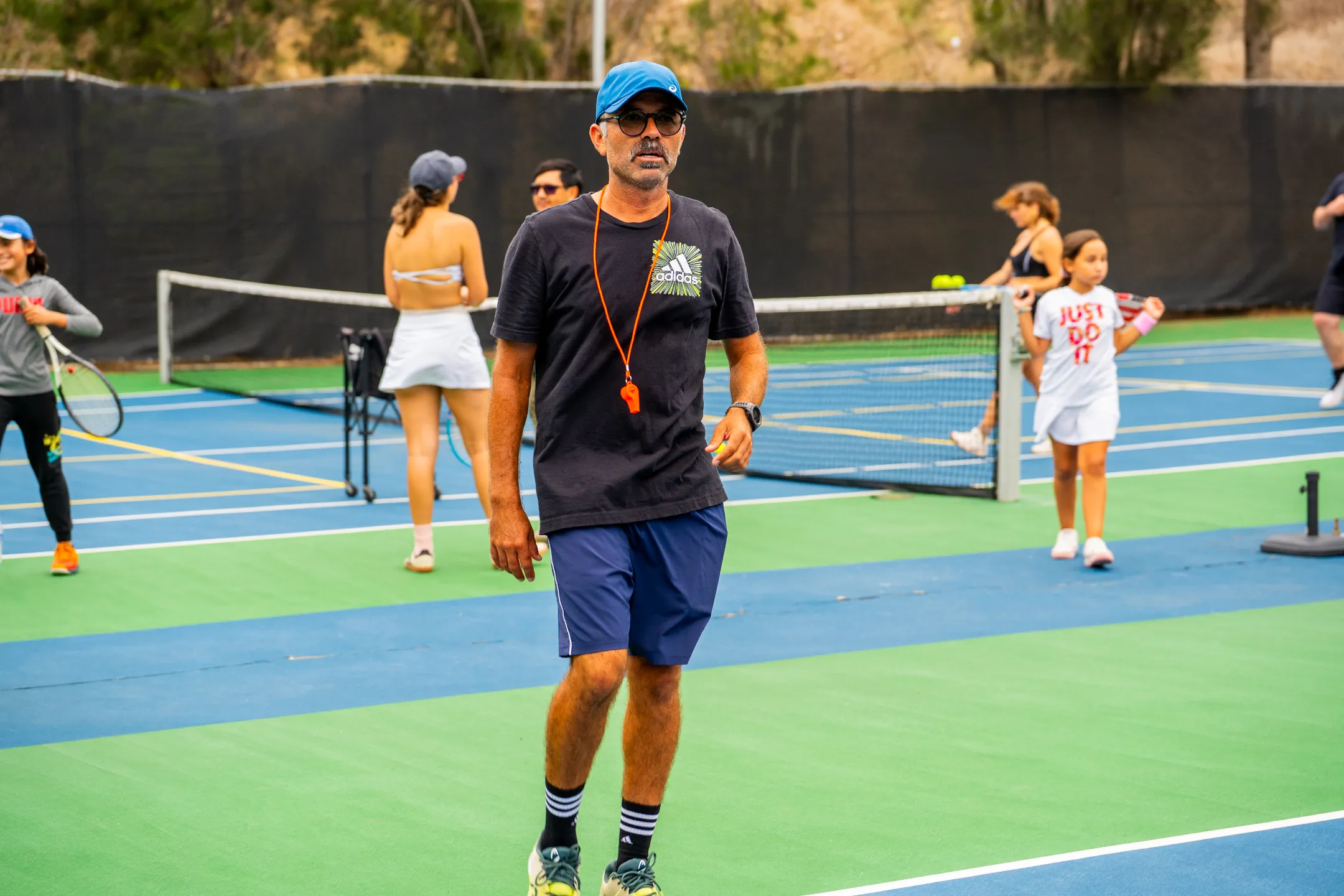 Entrenador con silbato en cancha de tenis mientras niños y jóvenes practican tenis.