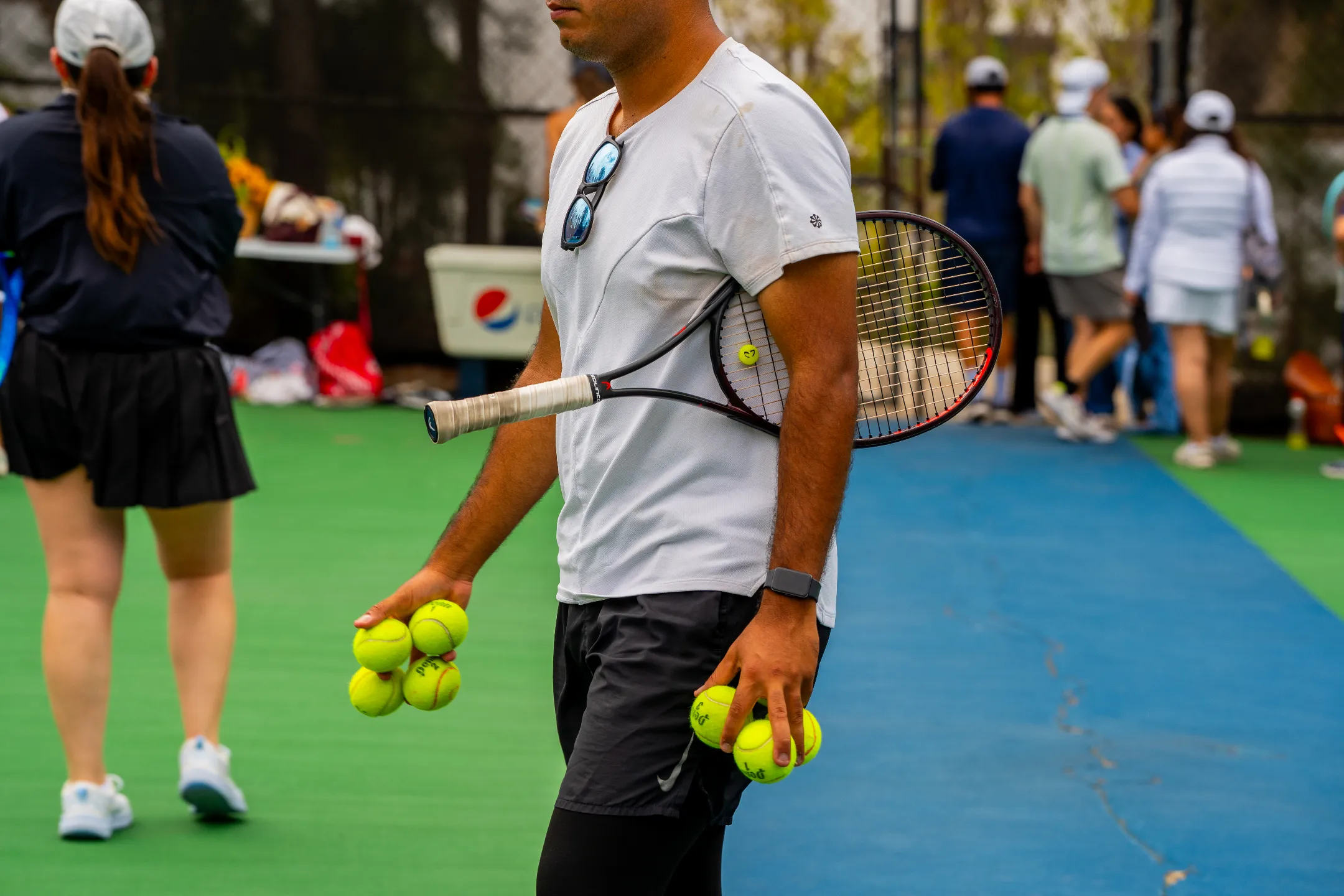 Hombre con raqueta y pelotas de tenis listas para jugar en cancha con personas al fondo.