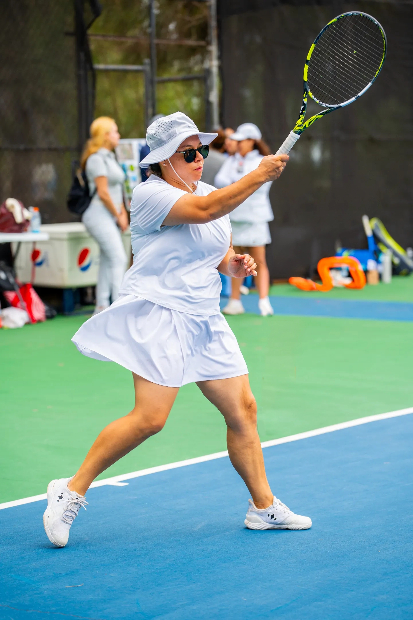 Mujer jugando tenis en cancha con vestido blanco, gorro y gafas oscuras, levantando raqueta.