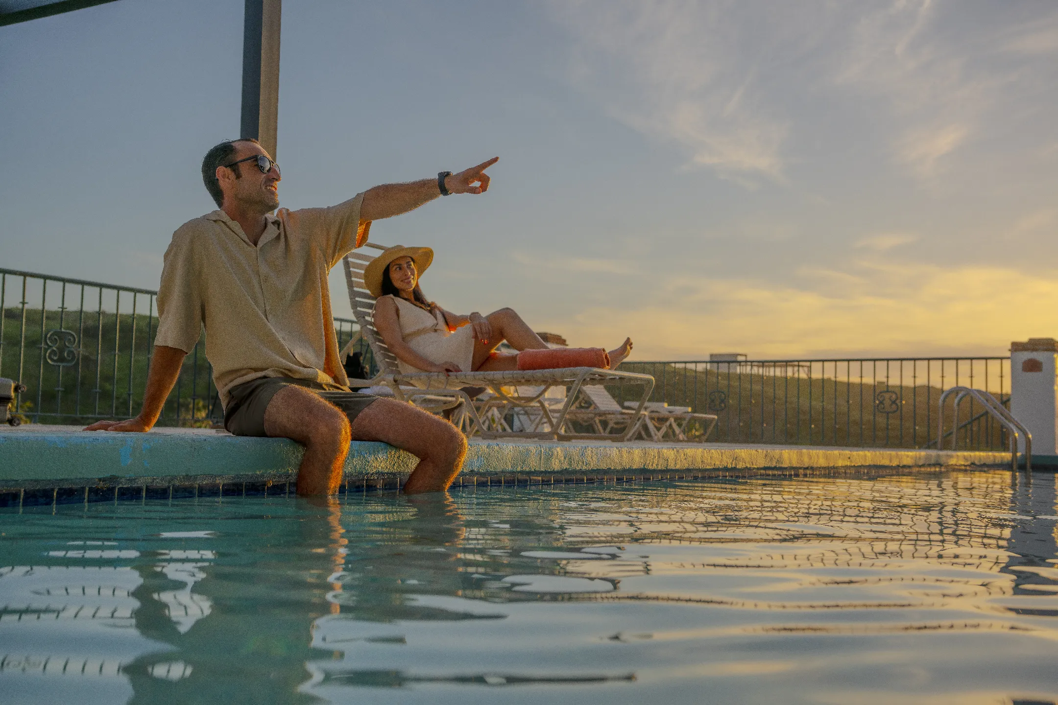 Hombre con gafas sentado al borde de la piscina y mujer recostada en una silla mientras cae el sol.