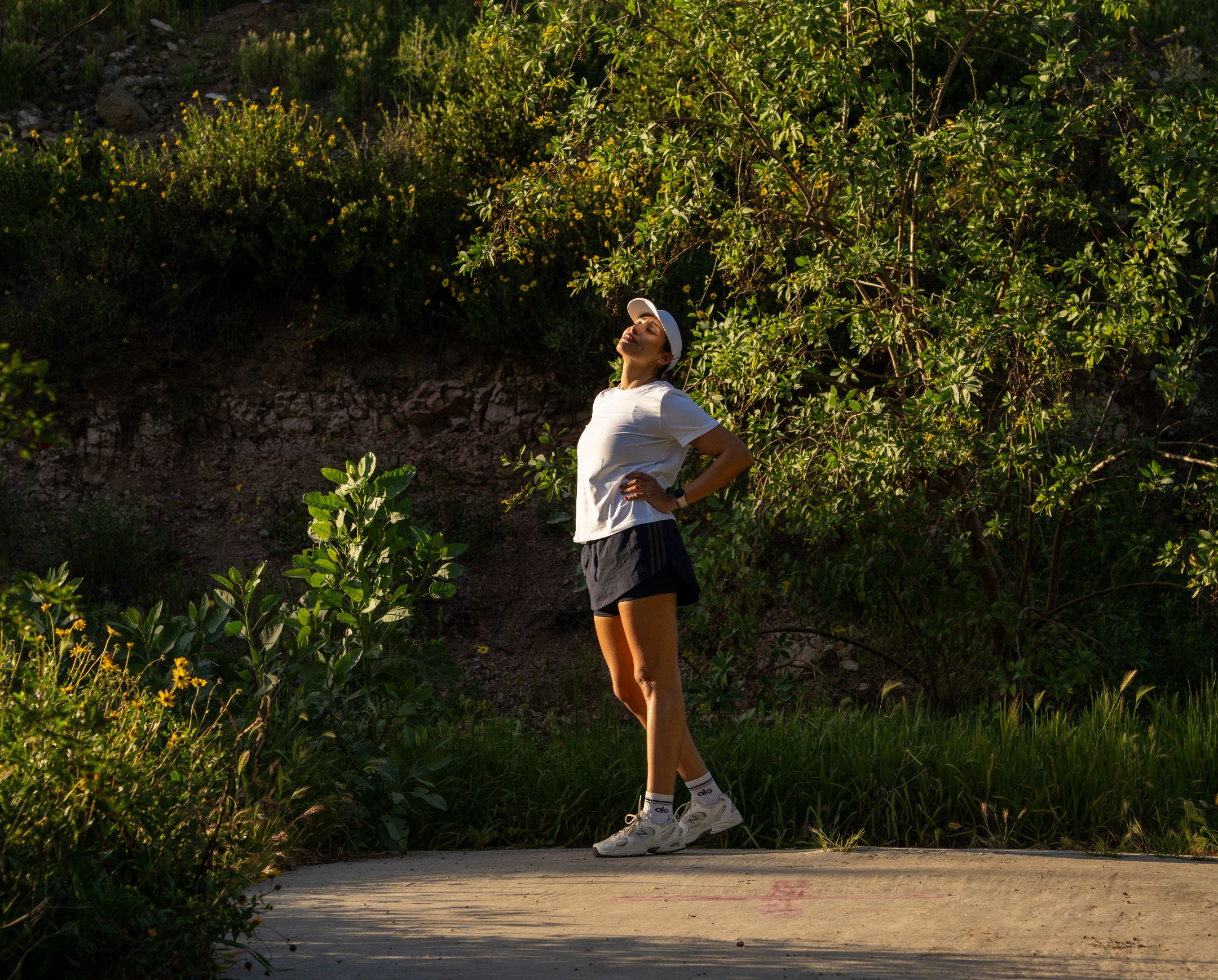 Mujer con gorra blanca y ropa deportiva disfrutando al aire libre en un sendero rodeado de vegetación.
