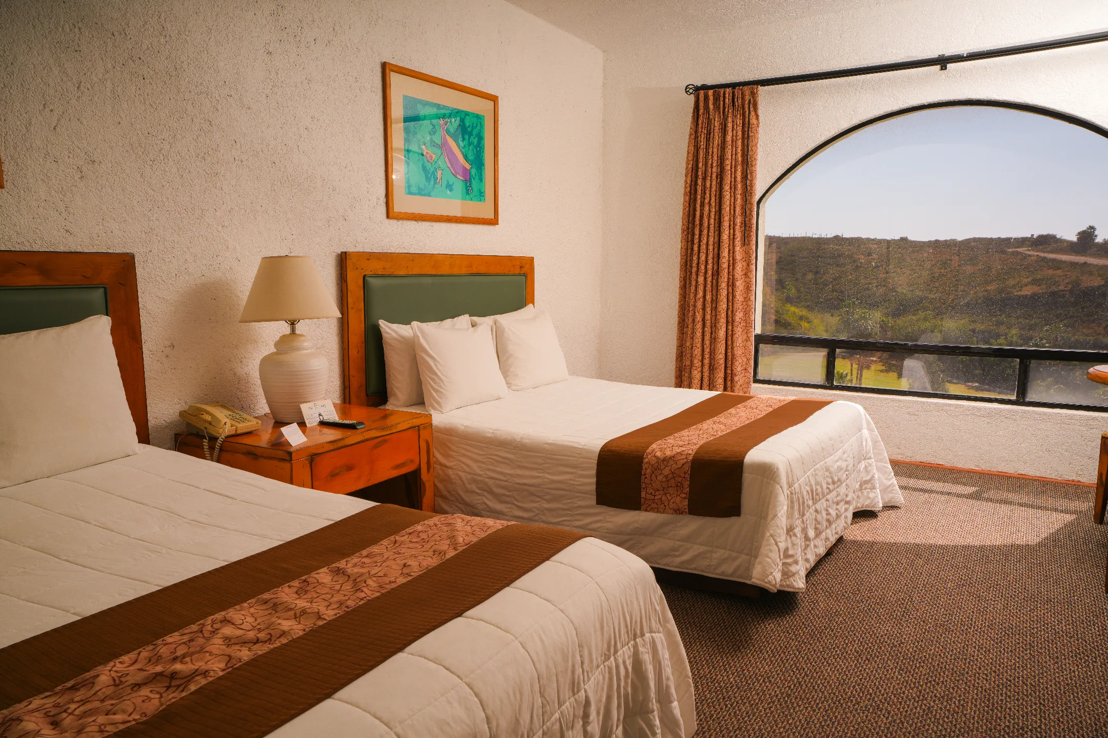 Habitación de hotel con dos camas, cabeceras de madera, lámpara, teléfono y ventana grande con vista al campo.