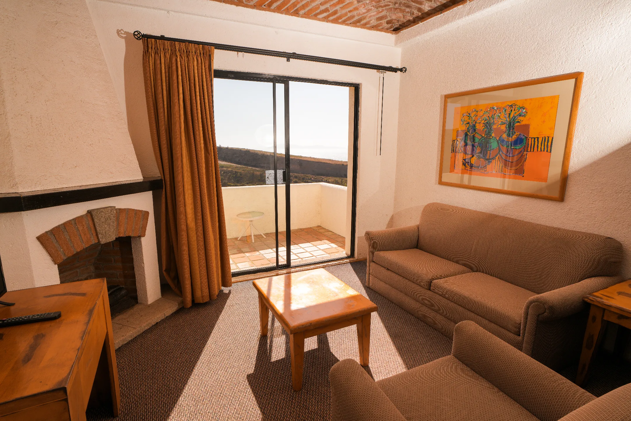 Sala con sofá, sillón, mesa, chimenea, puerta corrediza a balcón con vista de colinas.