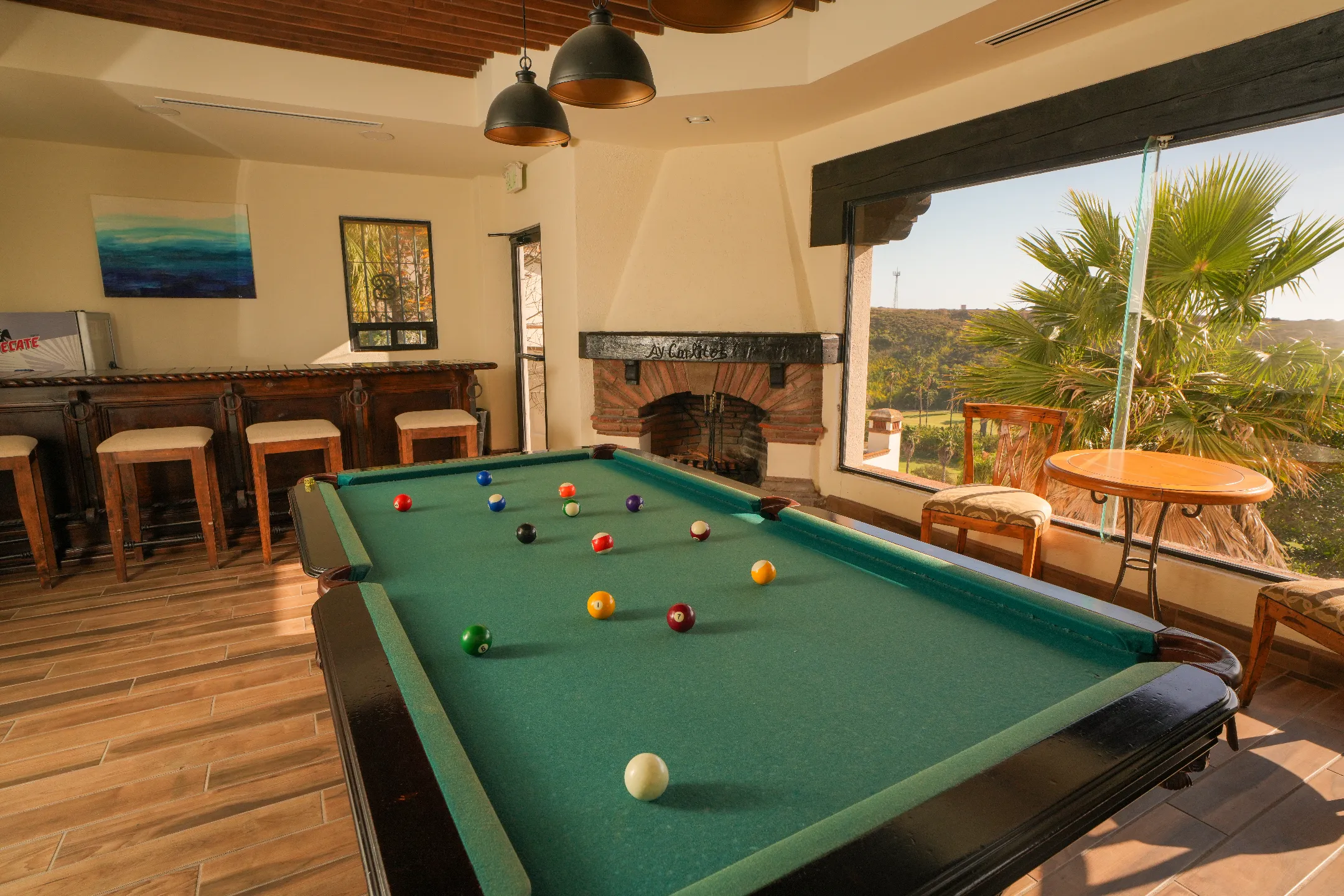 Mesa de billar con bolas en juego en habitación con chimenea, barra de madera y ventana con vista a palmeras.