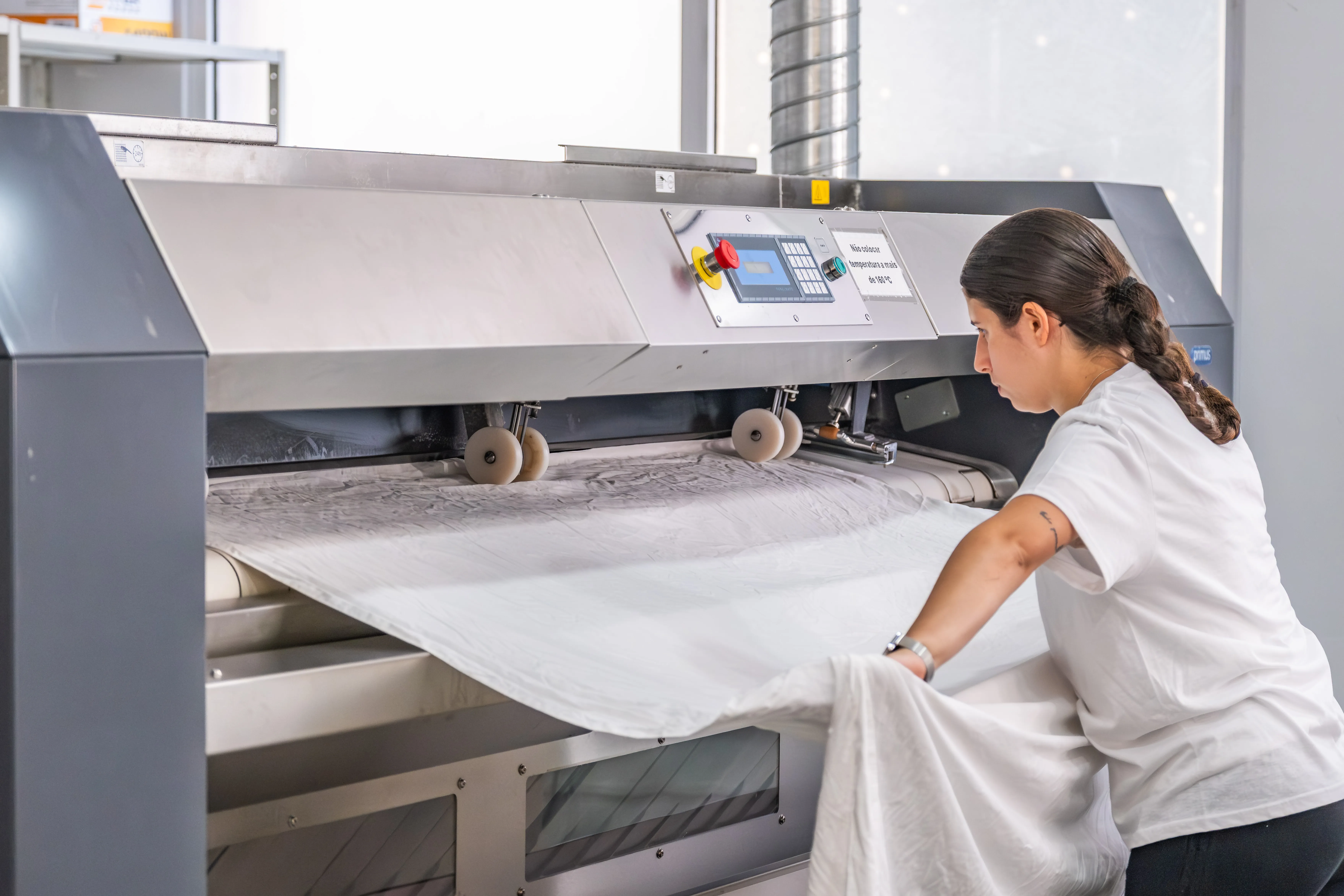 Processo de engomadoria industrial, com colaboradora a manusear têxteis profissionais em equipamento de precisão.