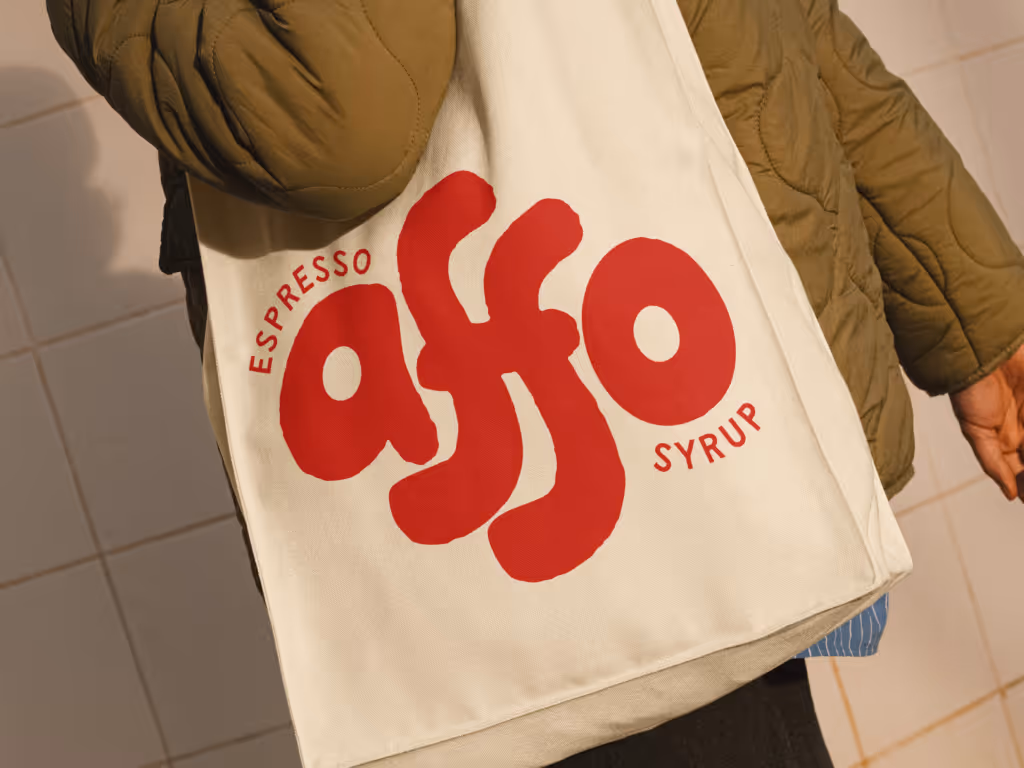 Person wearing an olive green jacket carrying a white tote bag with bold red text reading 'espresso affo syrup'.