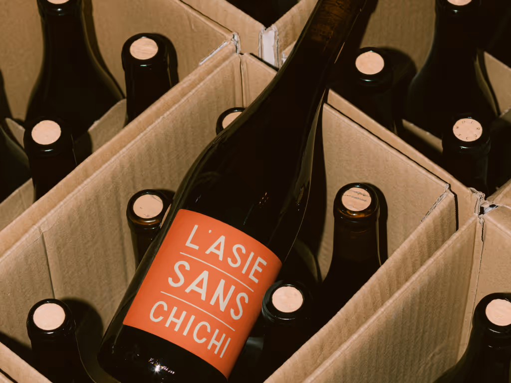 Dark wine bottle with an orange label reading 'L'ASIE SANS CHICHI' resting inside a cardboard box filled with other sealed bottles.