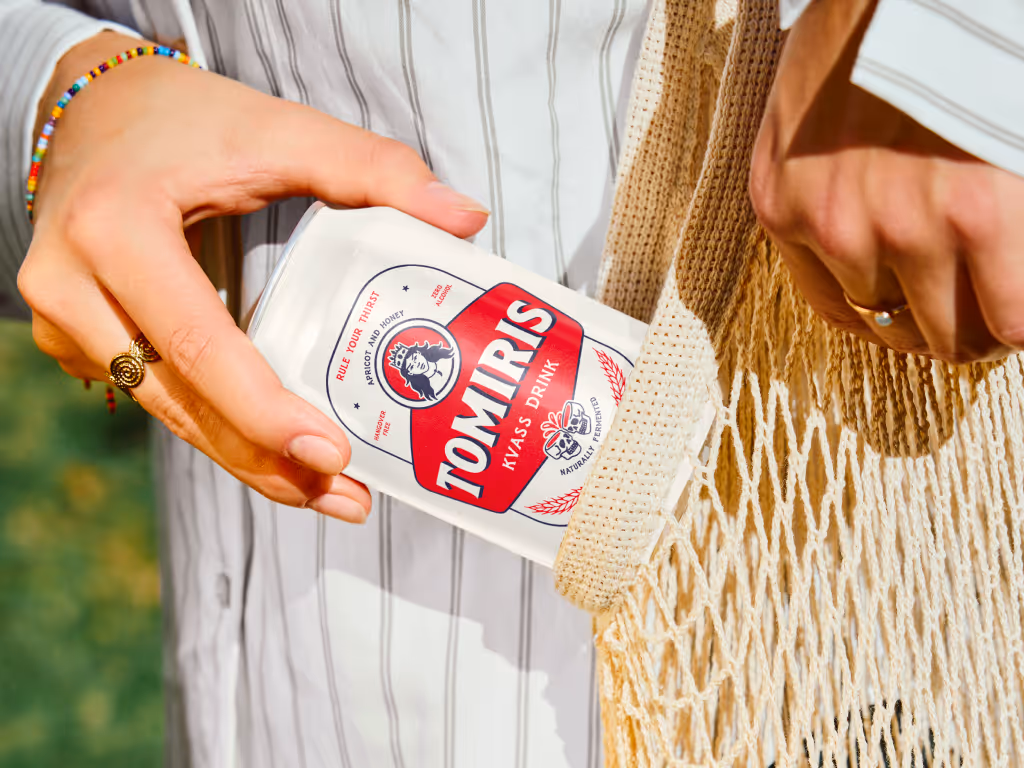 Close-up of a person’s hands holding a can of Tomiris kvass drink and placing it into a beige net shopping bag outdoors.