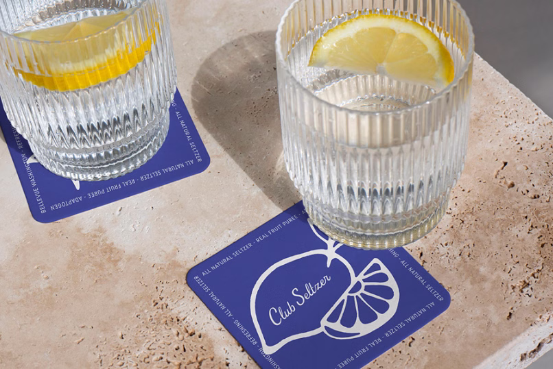 Two ribbed glasses each with a lemon slice and clear sparkling water on blue coasters labeled 'Club Seltzer' on a textured beige surface.