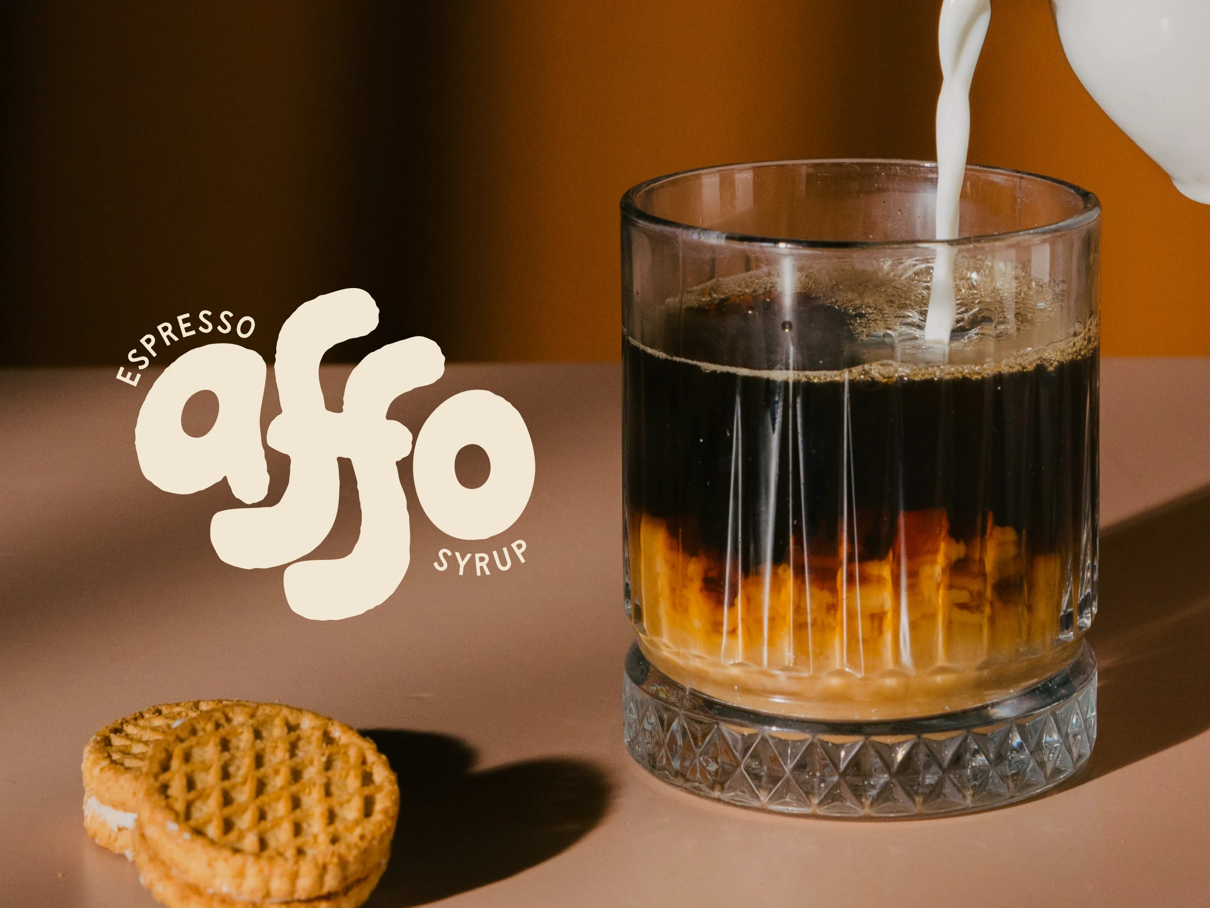 Glass of layered espresso drink with brown syrup at the bottom and dark espresso on top, milk being poured into it, with two round cream-filled cookies on the table and text 'espresso affo syrup' on the left.