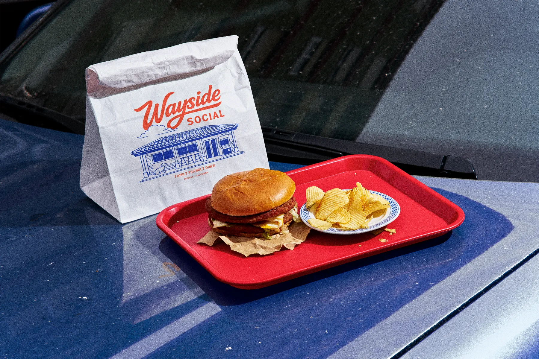 Red tray on a blue car hood holding a cheeseburger on brown paper and a small plate of wavy potato chips, with a white paper bag labeled 'Wayside Social Family Friendly Diner Moraga California' in the background.