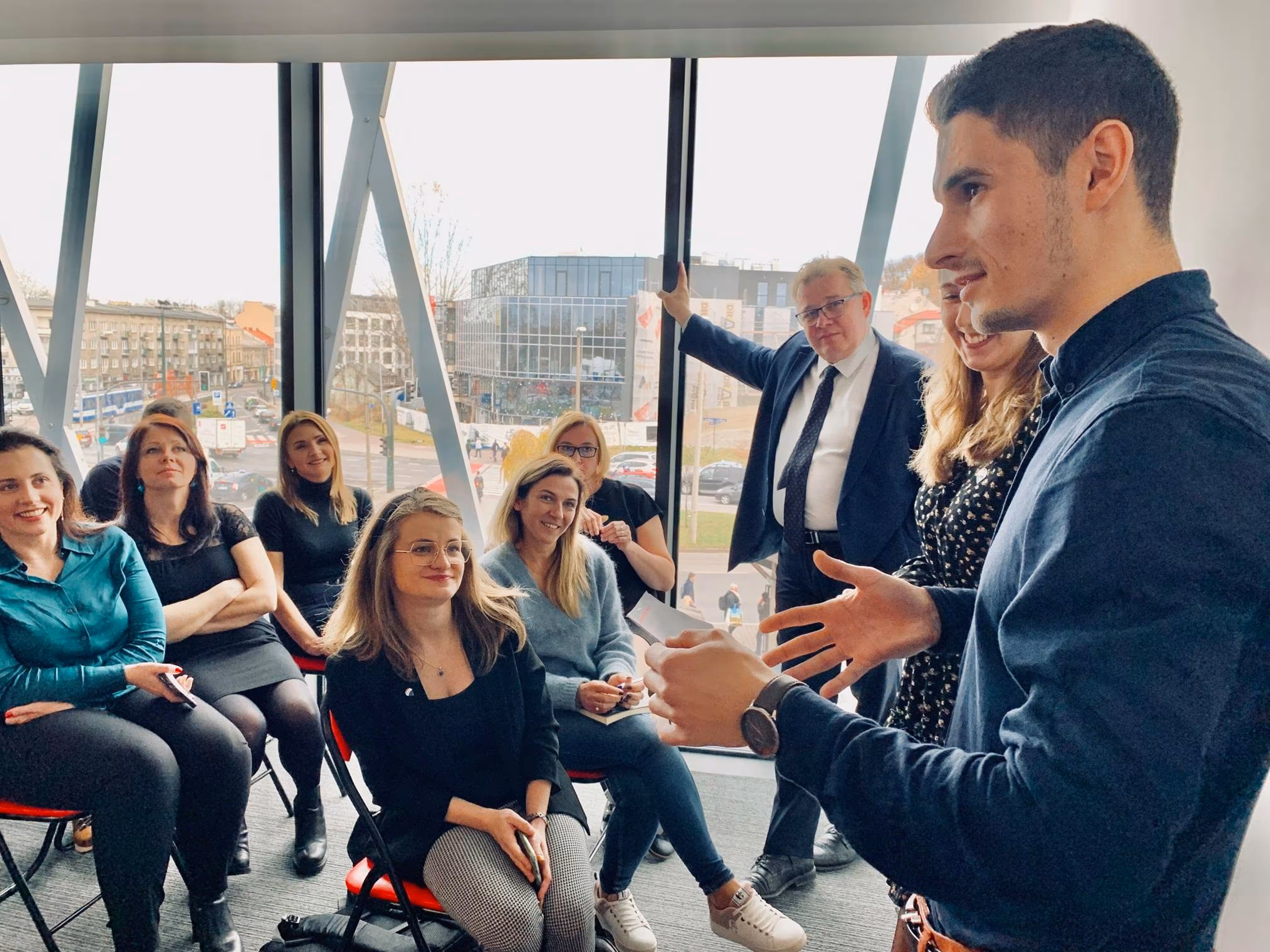 Młody mężczyzna przemawia do grupy osób siedzących na krzesłach w jasnym pokoju z dużymi oknami.