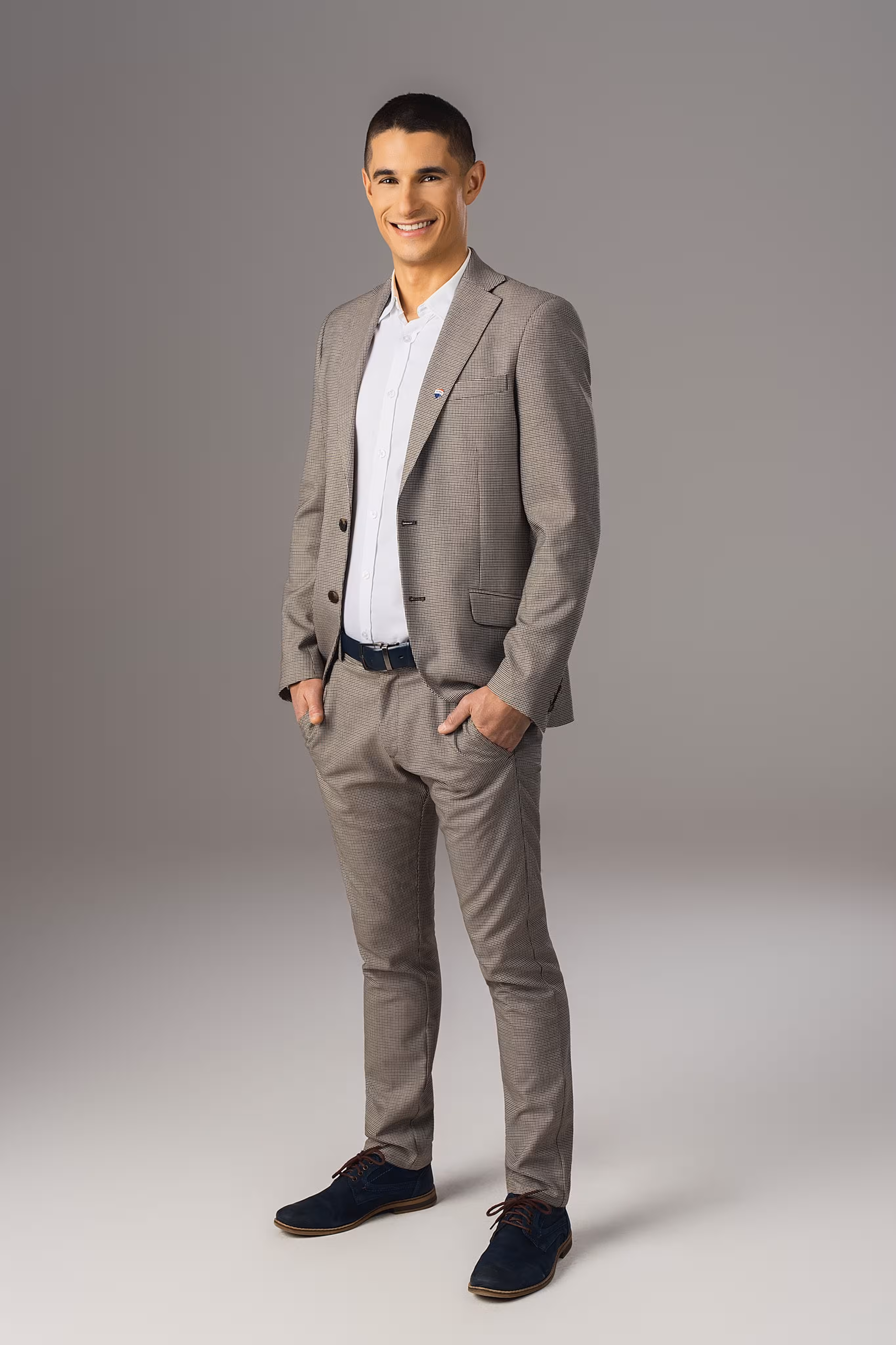 Maciej Tomalak w beżowym garniturze i białej koszuli, uśmiechający się ze włożonymi rękami do kieszeni, na szarym tle.