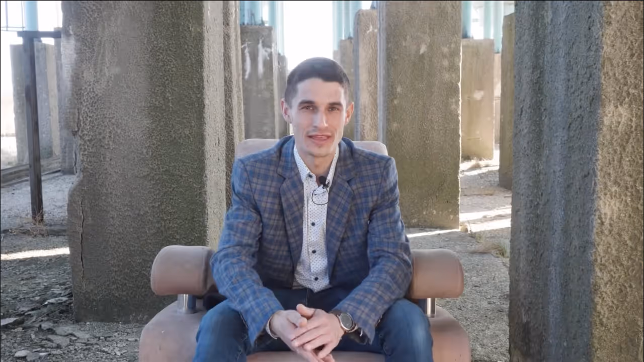 Młody mężczyzna w marynarce w kratę siedzi na fotelu w otoczeniu betonowych słupów.