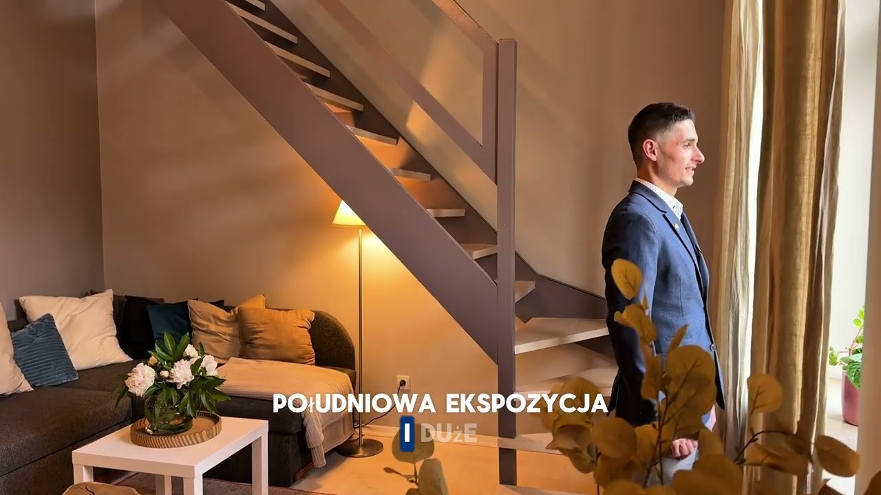 Mężczyzna w marynarce stoi przy oknie w nowocześnie urządzonym salonie ze schodami i sofą.