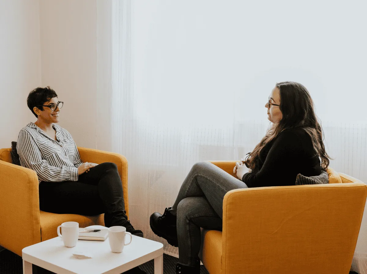 Dos mujeres conversan sentadas en sillones amarillos durante una sesión presencial, con dos tazas sobre la mesita