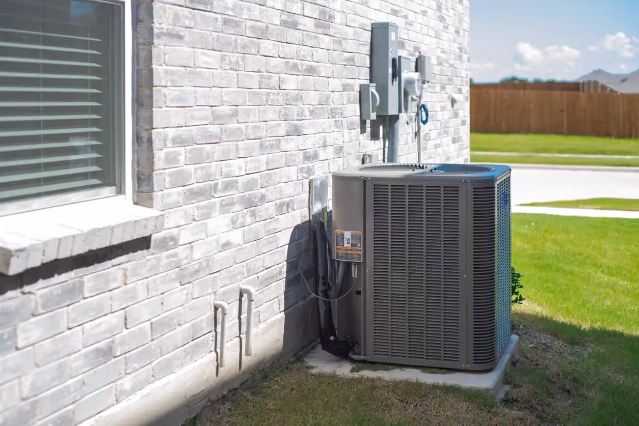 A dark grey heat pump condenser unit next to a light gray brick wall of a house. A window is visible on the left, and electrical components are mounted on the wall above the unit