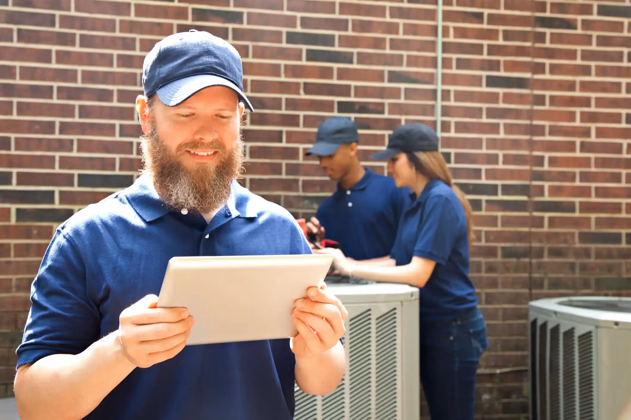 Smiling HVAC technician using a tablet while a team checks an outdoor air conditioning unit.