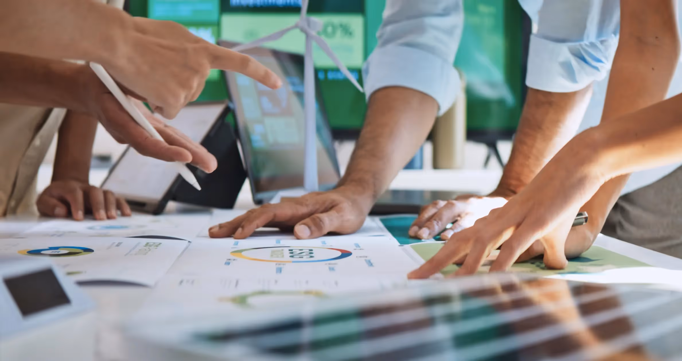 A group of people huddle around a table covered in architectural plans and charts, discussing energy-efficient designs with a wind turbine model visible in the background.