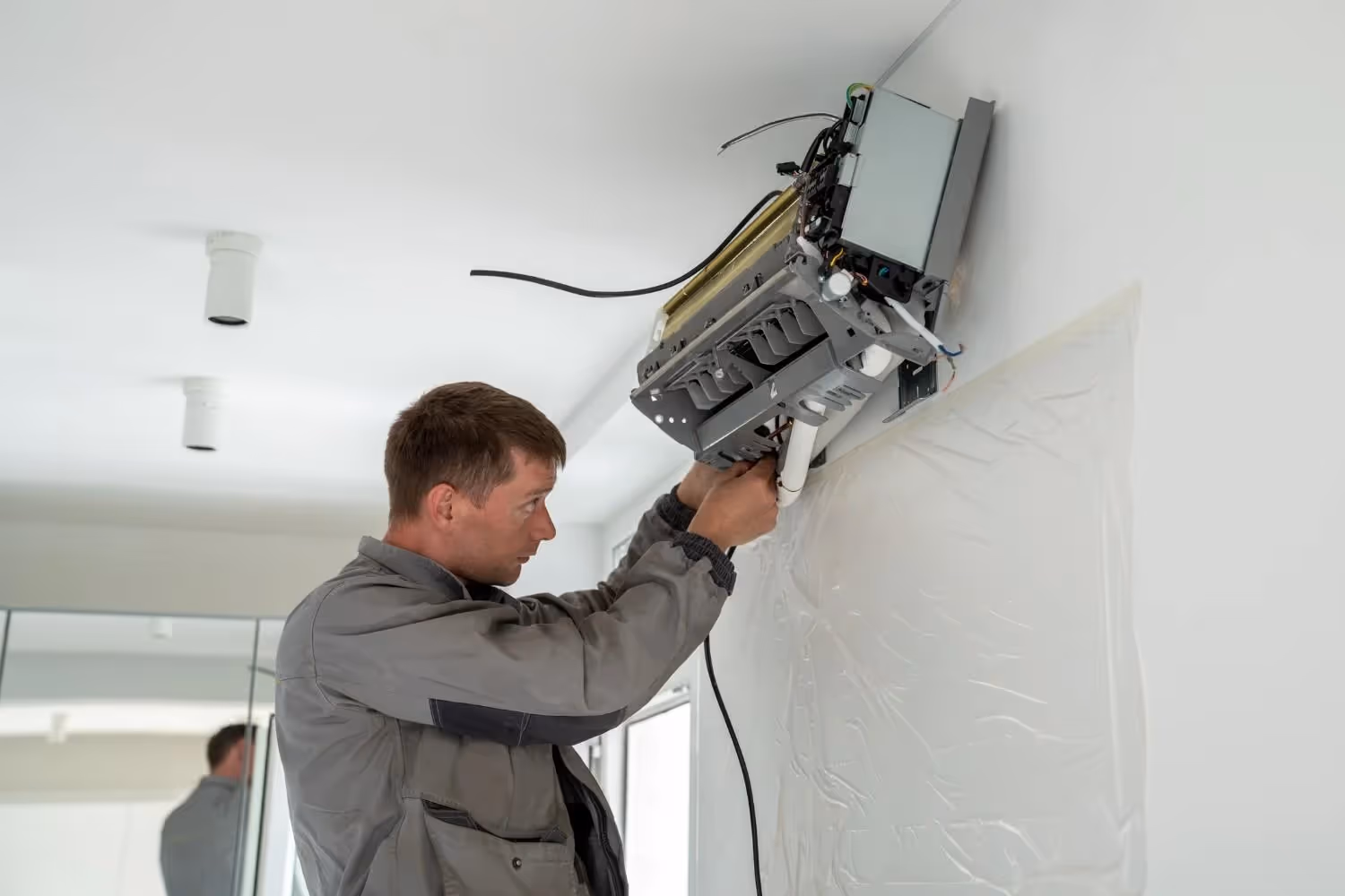 A technician in a gray work jacket performing maintenance or installation on a wall-mounted air conditioning unit. The unit’s casing is open, exposing internal electrical components and wiring. A protective plastic sheet is taped to the wall below the unit, and the indoor setting features ceiling lights and a mirror reflecting part of the room.