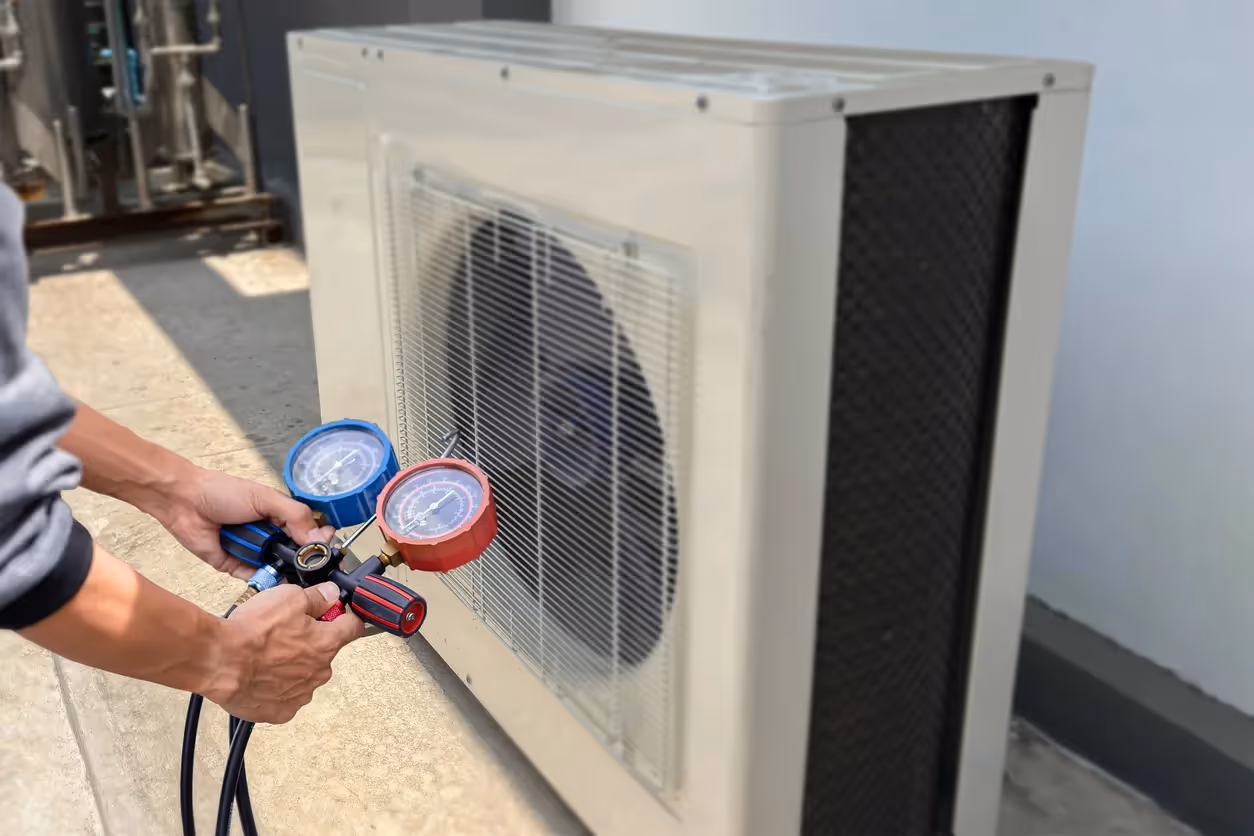 A technician is holding a manifold gauge set with red and blue gauges and hoses, performing a check on a modern, beige-colored outdoor HVAC unit on a sunny rooftop. The unit has a protective grille over the fan.