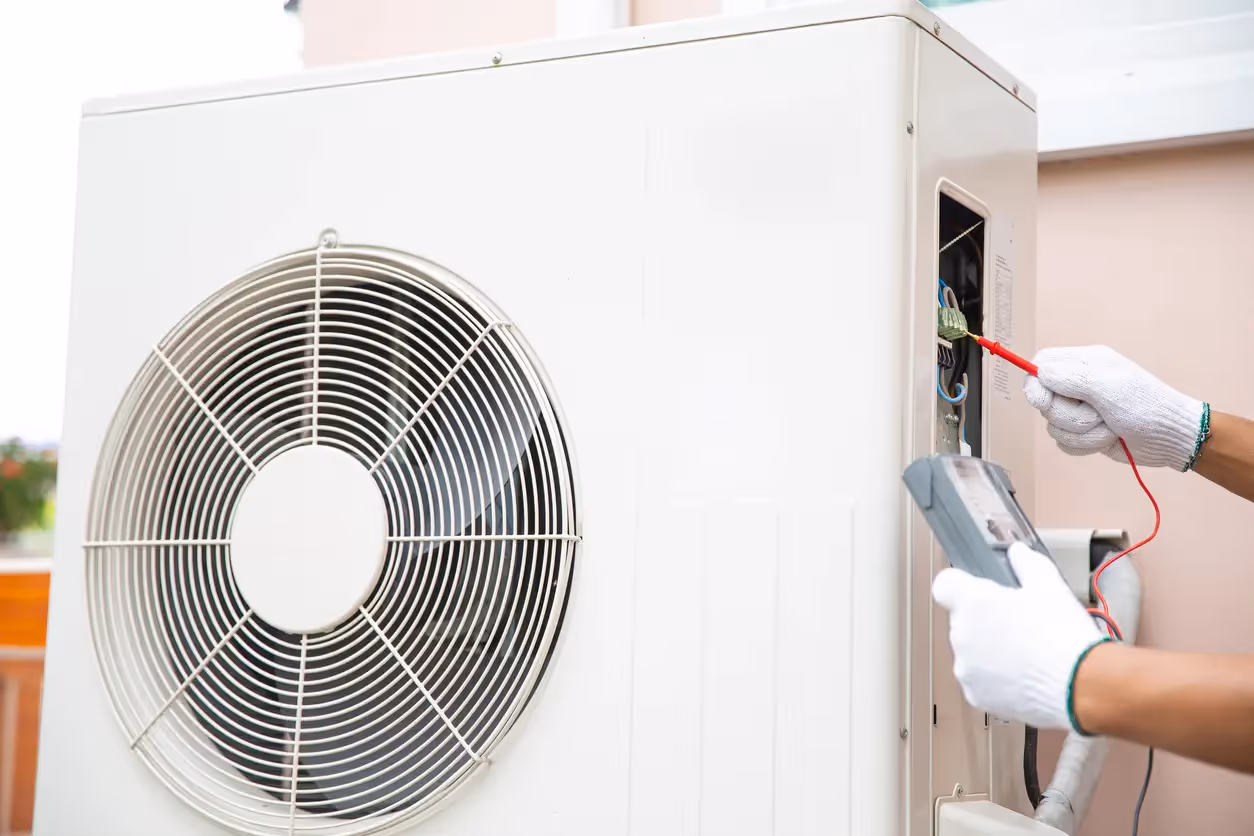 A close-up of a white outdoor HVAC unit shows a technician in white gloves using a multimeter to check electrical connections inside the service compartment.