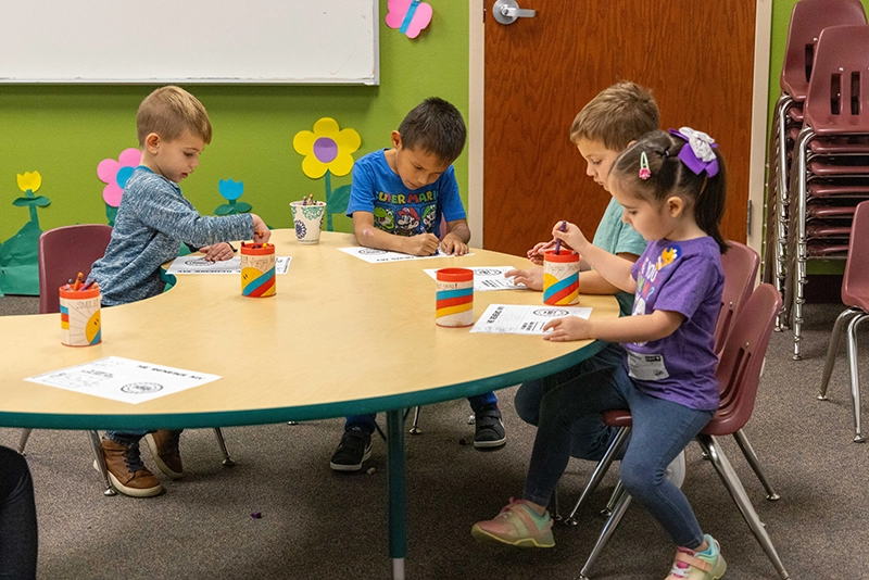 Four young children sitting around a table coloring on worksheets with crayons in a classroom decorated with colorful paper flowers. Children's ministry West Valley Church.