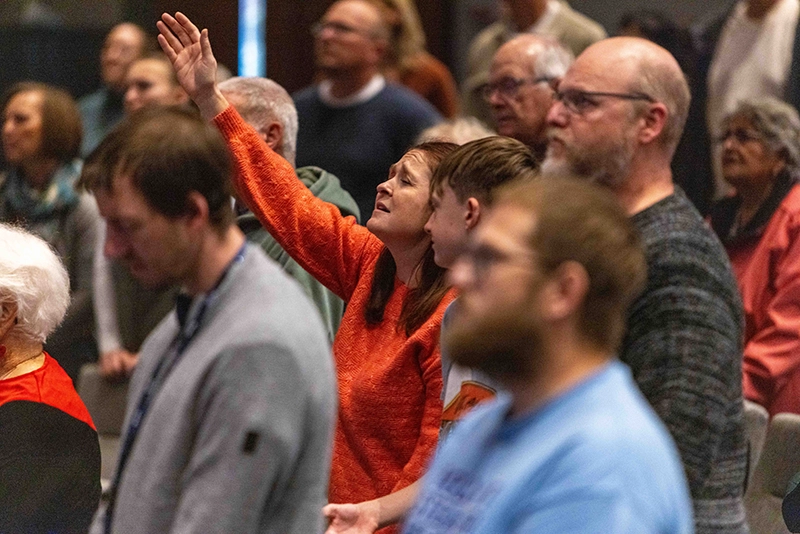 Woman in an orange sweater with her hand raised, surrounded by a diverse group of people standing in the worship center at West Valley Church.