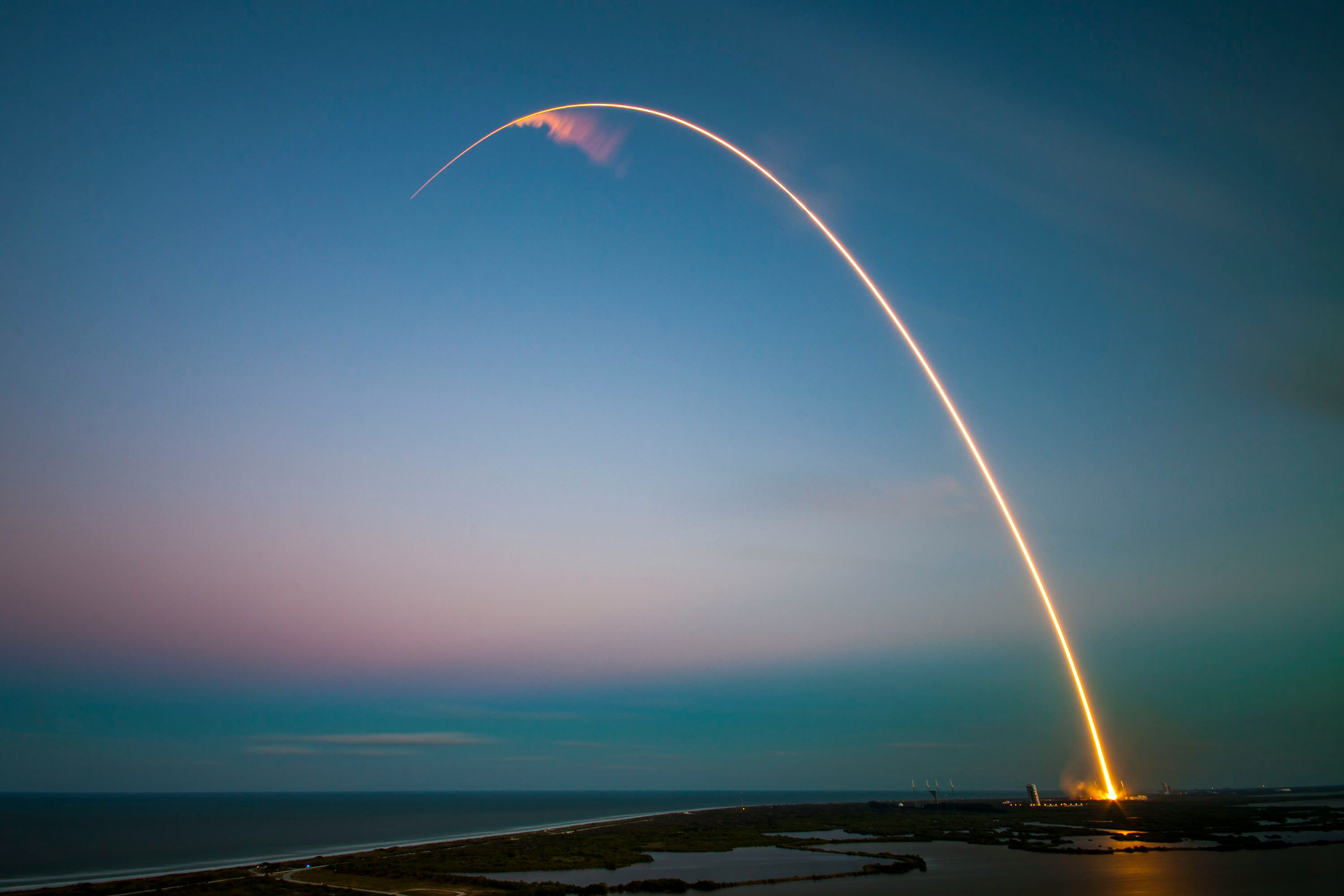 Long exposure of a rocket launch showing a bright arc of light against a dusk sky over a coastal landscape.