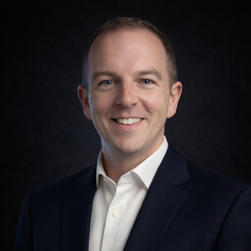 Smiling man in a dark suit and white shirt against a black background.