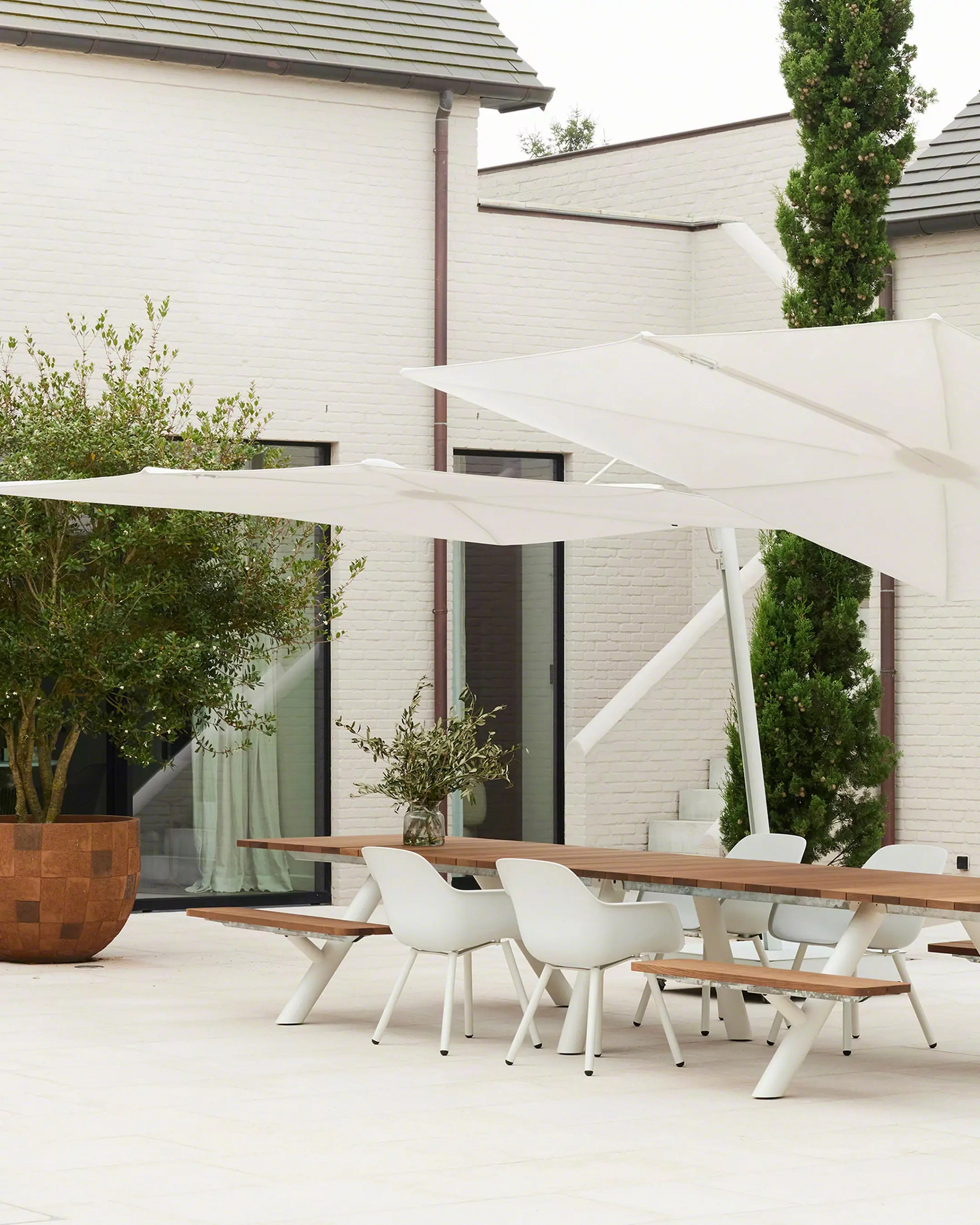 Modern living room with a curved off-white sofa and matching pillow, beside a black side table with metallic angled lamps, large windows showing greenery outside.